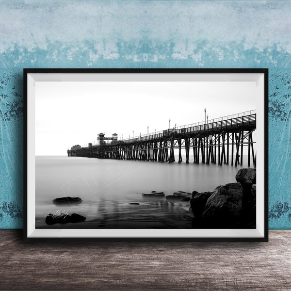 A black and white photograph of a long pier extending into the water, with a rocky shoreline in the foreground.