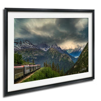 A framed photograph depicting a train traveling through a mountainous landscape with snow-capped peaks and a dramatic cloudy sky.