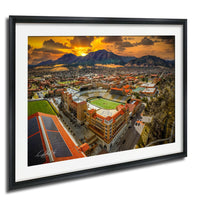 The image is a framed aerial view of a city with a large stadium, mountains in the background, and a dramatic sunset sky.