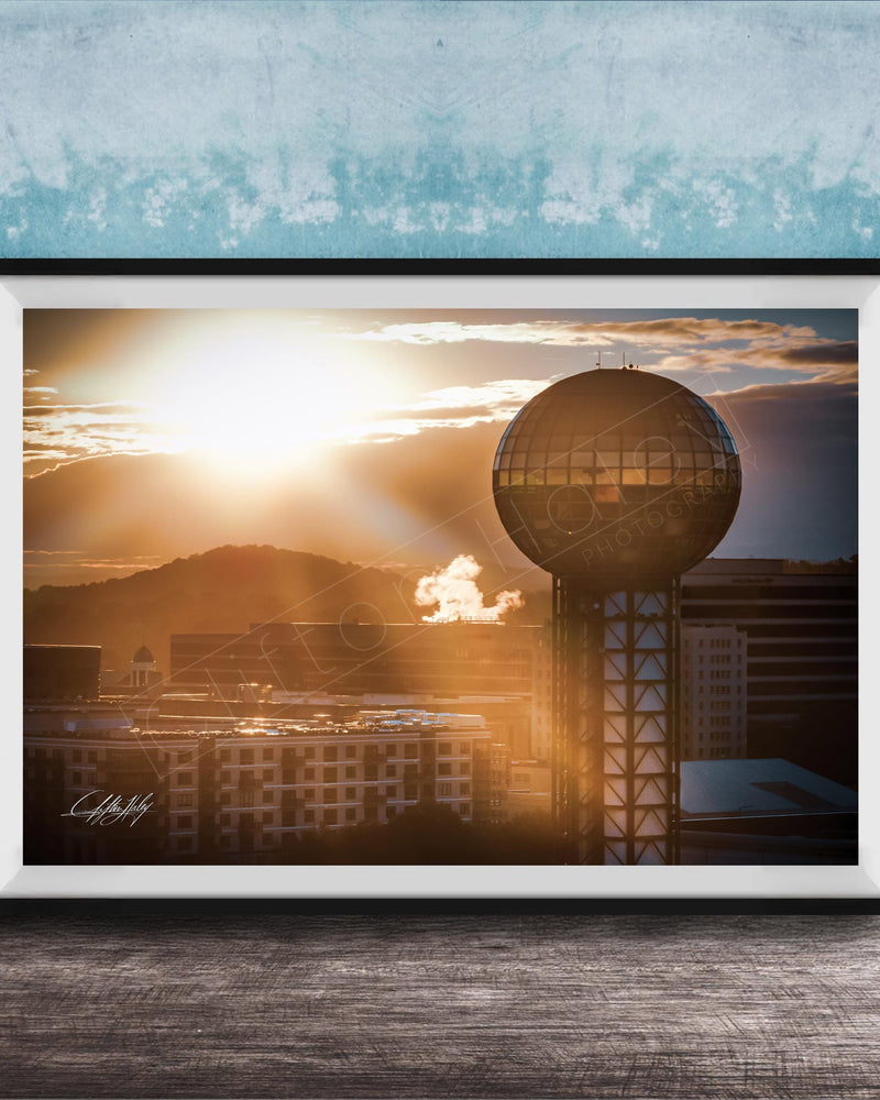 A framed photograph of a city skyline at sunset, with a large tower in the foreground and the sun setting behind the buildings.