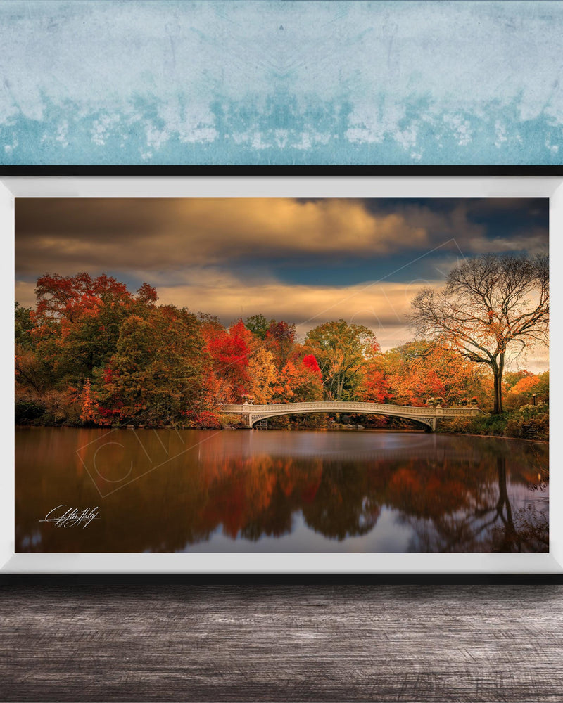 A framed photograph depicting a scenic autumn landscape with a bridge, trees, and a body of water reflecting the vibrant colors of the season.