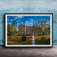 A framed photograph of a brick building with a tower, surrounded by trees and a grassy area, set against a blue sky with clouds.