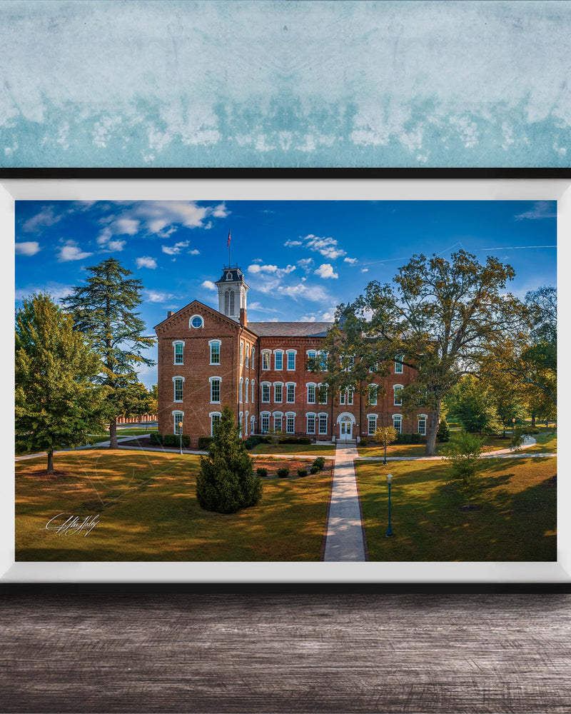 A framed photograph of a brick building with a tower, surrounded by trees and a grassy area, set against a blue sky with clouds.