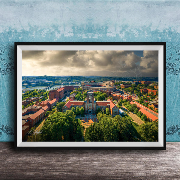 The image is a framed aerial view of a city with a mix of buildings, trees, and a river. The sky is cloudy, creating a dramatic backdrop for the cityscape.