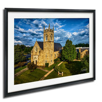 A stone church with a tall tower and a clock, surrounded by trees and a grassy area, set against a blue sky with clouds.