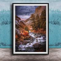 The image is a framed photograph of a scenic mountain landscape with a winding river flowing through it. The mountains are covered in snow, and the sky is filled with clouds, creating a dramatic and moody atmosphere. The photograph is framed and displayed on a wooden surface, with a blue wall in the background.
