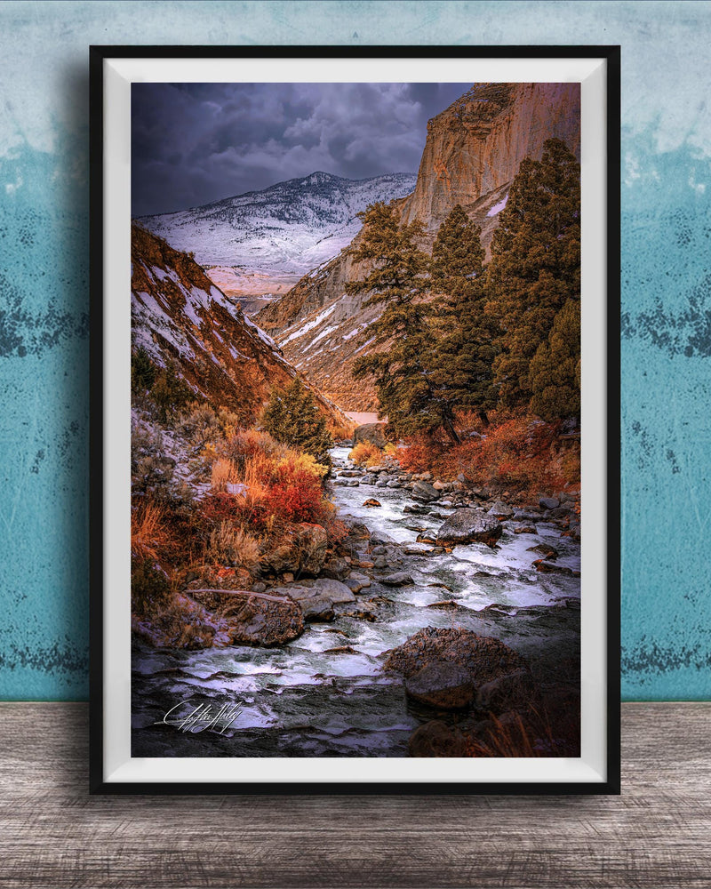The image is a framed photograph of a scenic mountain landscape with a winding river flowing through it. The mountains are covered in snow, and the sky is filled with clouds, creating a dramatic and moody atmosphere. The photograph is framed and displayed on a wooden surface, with a blue wall in the background.