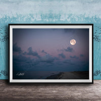A framed photograph of a serene beach at night, with a full moon rising over the ocean and a cloudy sky.