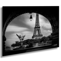 A black and white photograph of the Eiffel Tower in Paris, France, with a statue of a winged figure in the foreground and a stone archway framing the scene.