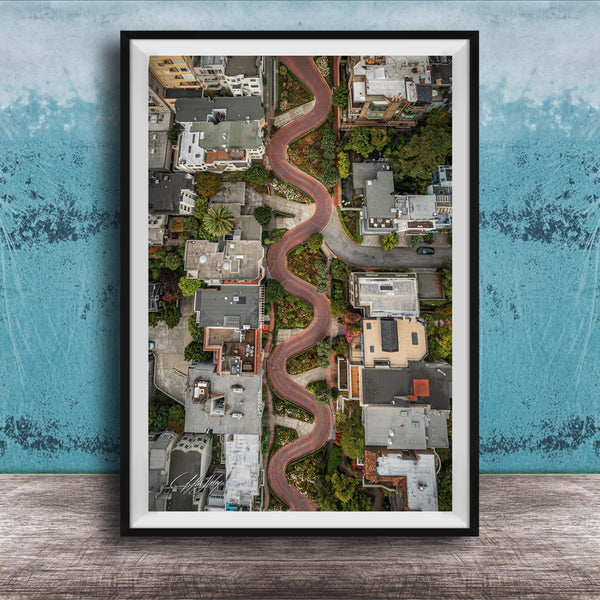 The image is an aerial view of a winding road that curves through a cityscape, with buildings and greenery lining the sides of the road.