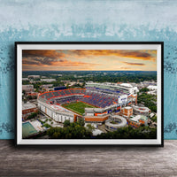 The image is a framed photograph of a large sports stadium, likely a football stadium, with a colorful sunset sky in the background.