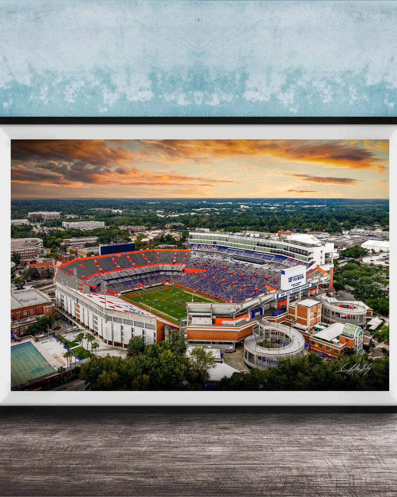 The image is a framed photograph of a large sports stadium, likely a football stadium, with a colorful sunset sky in the background.