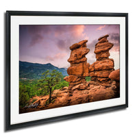 A framed photograph depicting a scenic landscape with two towering rock formations, possibly rock stacks or sandstone pillars, set against a backdrop of mountains and a colorful sky.