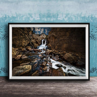 A framed photograph of a rocky waterfall cascading over rocks, set against a blue wall with a textured pattern.