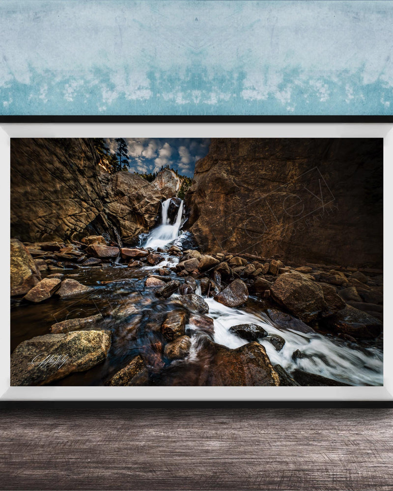A framed photograph of a rocky waterfall cascading over rocks, set against a blue wall with a textured pattern.