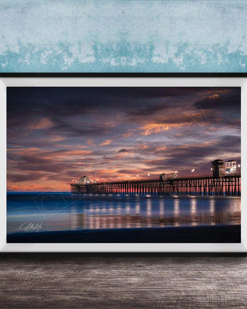 A framed photograph of a wooden pier extending into the ocean at sunset, with the sky filled with vibrant colors and clouds.