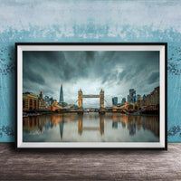 A framed photograph of the Tower Bridge in London, England, with the city's skyline reflected in the water below. The sky is filled with clouds, creating a dramatic atmosphere.