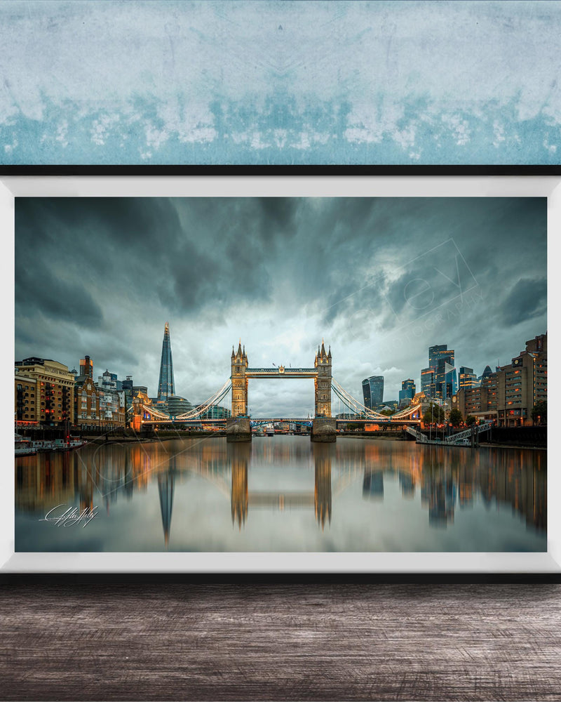 A framed photograph of the Tower Bridge in London, England, with the city's skyline reflected in the water below. The sky is filled with clouds, creating a dramatic atmosphere.