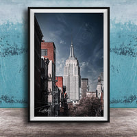 A framed photograph of the iconic Empire State Building in New York City, with a blue sky and clouds in the background.