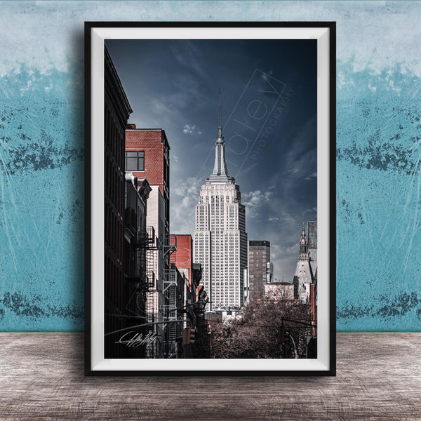 A framed photograph of the iconic Empire State Building in New York City, with a blue sky and clouds in the background.