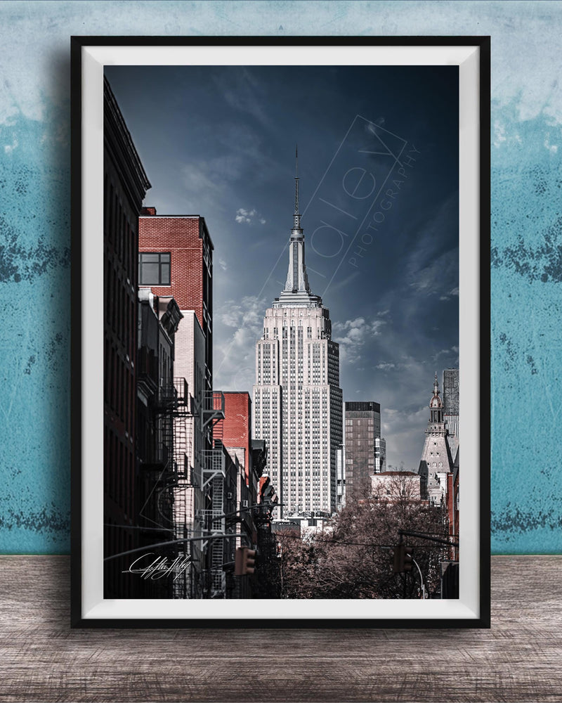 A framed photograph of the iconic Empire State Building in New York City, with a blue sky and clouds in the background.