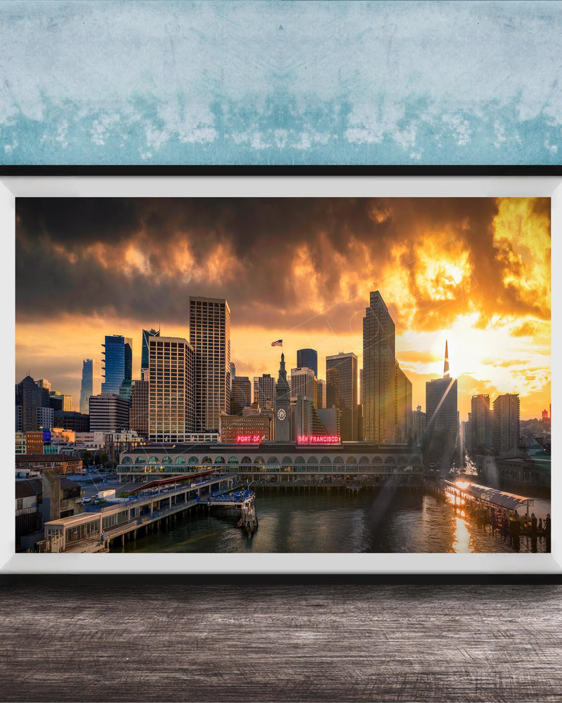 A framed photograph of a city skyline at sunset, with the sun setting behind the buildings and casting a warm, golden glow over the scene.