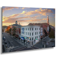 a city street scene with a mix of architectural styles, including a tall white building with a blue sign and a red brick building with a clock tower. The sky is filled with clouds, and the sun is setting, casting a warm glow over the scene.