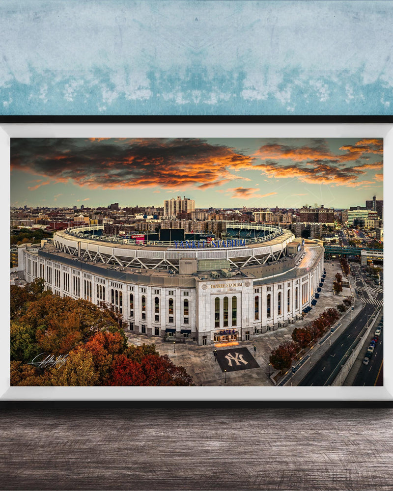 The image is a framed photograph of the New York Yankees stadium, which is a large, circular stadium located in the city of New York. The photograph captures the stadium's distinctive architecture and surrounding cityscape, with a dramatic sky in the background.