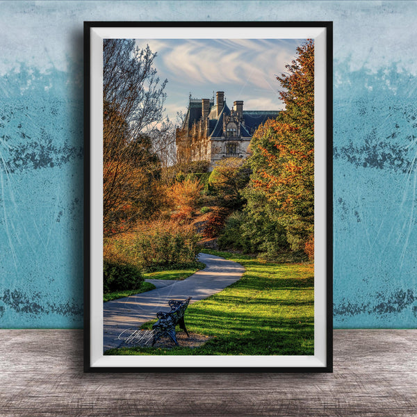 A framed photograph of a park with a winding path, a bench, and trees with autumn foliage. In the background, there is a large, ornate building with a gabled roof and multiple chimneys.