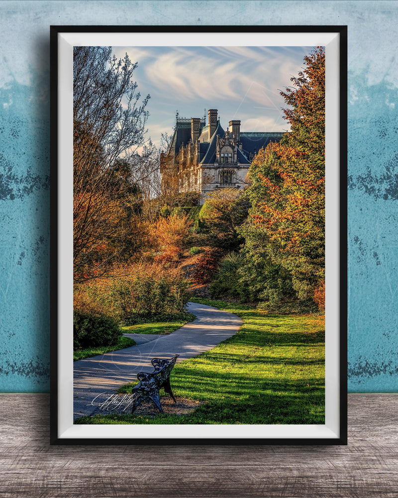 A framed photograph of a park with a winding path, a bench, and trees with autumn foliage. In the background, there is a large, ornate building with a gabled roof and multiple chimneys.