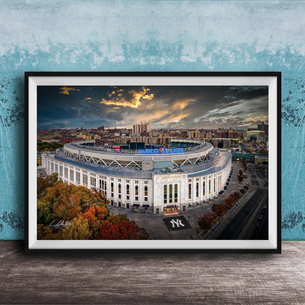 The image is a framed photograph of the New York Yankees stadium, which is a large, circular stadium located in the city of New York. The photograph captures the stadium's distinctive architecture and surrounding cityscape, with a dramatic sky in the background.