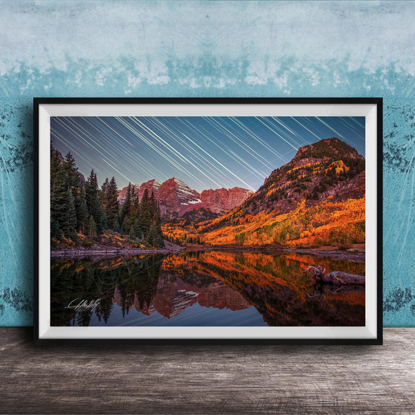 The image is a framed photograph depicting a serene mountain landscape with a lake reflecting the vibrant colors of the autumn foliage. The sky is filled with long exposure star trails, creating a stunning visual effect that adds depth and dimension to the scene.