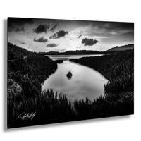 A black and white landscape photograph depicting a serene lake surrounded by a forested area, with mountains visible in the distance under a cloudy sky.