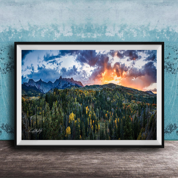 The image is a framed landscape photograph depicting a mountainous, forested area with a dramatic sky at sunset.