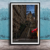 a city street with a tall, ornate building on a steep, winding staircase. The building has a distinctive dome and is surrounded by other buildings. The street is lined with trees and has outdoor seating areas, suggesting a cafe or restaurant. The sky is dark and cloudy, creating a moody atmosphere. The image is framed and displayed on a wooden surface, with a blue wall in the background.