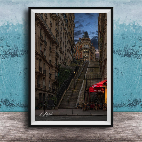 a city street with a tall, ornate building on a steep, winding staircase. The building has a distinctive dome and is surrounded by other buildings. The street is lined with trees and has outdoor seating areas, suggesting a cafe or restaurant. The sky is dark and cloudy, creating a moody atmosphere. The image is framed and displayed on a wooden surface, with a blue wall in the background.