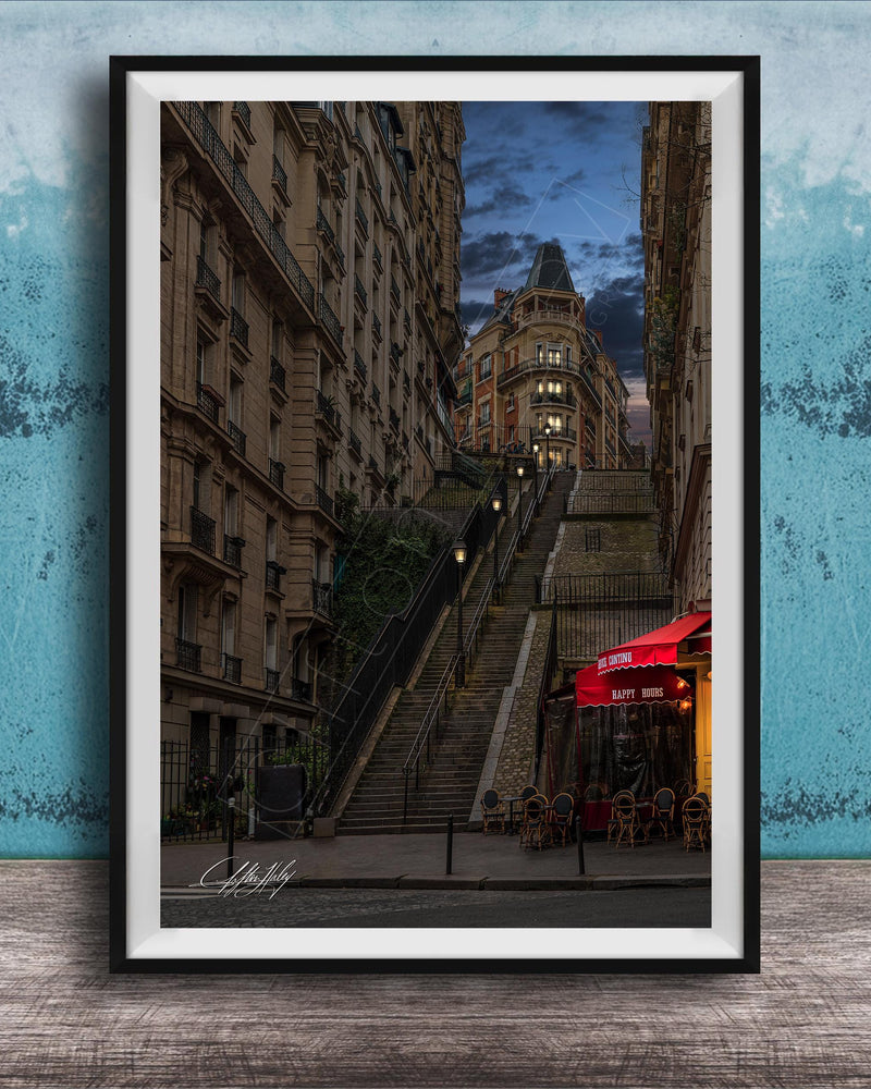 a city street with a tall, ornate building on a steep, winding staircase. The building has a distinctive dome and is surrounded by other buildings. The street is lined with trees and has outdoor seating areas, suggesting a cafe or restaurant. The sky is dark and cloudy, creating a moody atmosphere. The image is framed and displayed on a wooden surface, with a blue wall in the background.