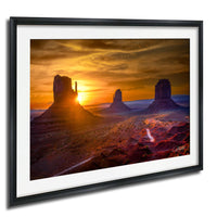 A framed photograph depicting the iconic rock formations of Monument Valley at sunset, with the sun setting behind the mountains and casting a warm, golden glow over the landscape.