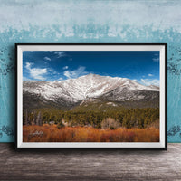 A framed photograph of a snow-capped mountain range with a forested valley in the foreground, set against a blue sky with wispy clouds.