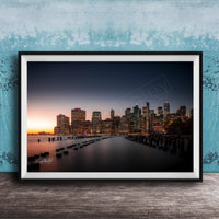 A framed photograph of the skyline of New York City at sunset, with the city's skyscrapers and landmarks illuminated against a dark blue sky.