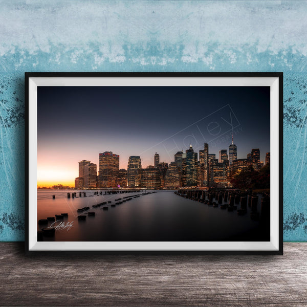 A framed photograph of the skyline of New York City at sunset, with the city's skyscrapers and landmarks illuminated against a dark blue sky.