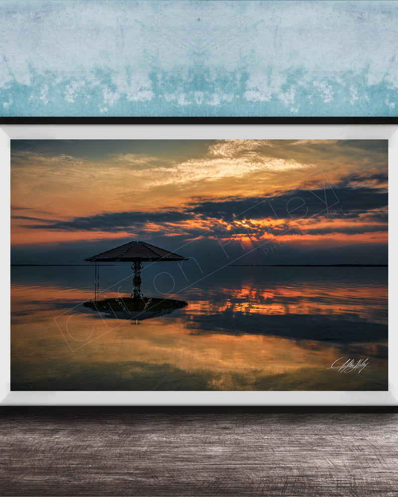 A framed photograph depicting a serene sunset over a calm body of water, with a lone umbrella standing in the foreground.
