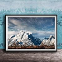A framed photograph of a snow-capped mountain range against a cloudy sky, displayed on a wooden surface against a blue textured wall.
