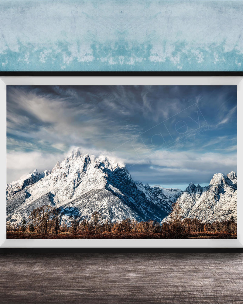 A framed photograph of a snow-capped mountain range against a cloudy sky, displayed on a wooden surface against a blue textured wall.