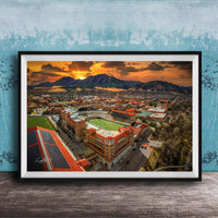The image is a framed aerial view of a city with a large stadium, mountains in the background, and a dramatic sunset sky.