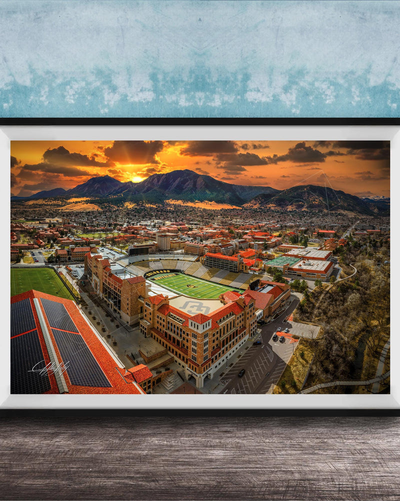 The image is a framed aerial view of a city with a large stadium, mountains in the background, and a dramatic sunset sky.