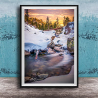 A framed photograph depicting a serene winter landscape with a snow-covered mountain, a flowing stream, and a vibrant sunset sky.