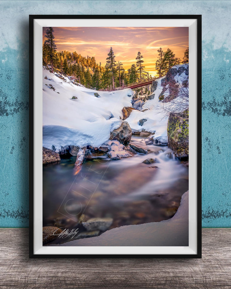A framed photograph depicting a serene winter landscape with a snow-covered mountain, a flowing stream, and a vibrant sunset sky.