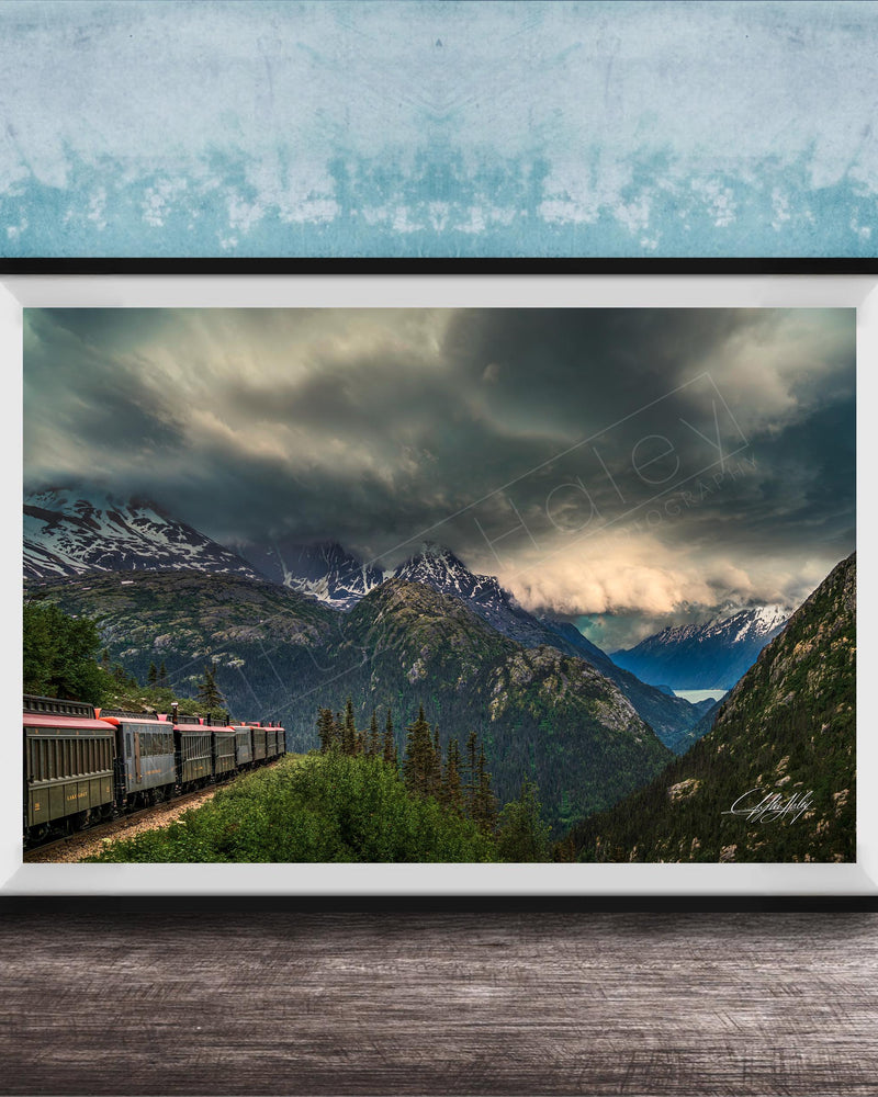 A framed photograph depicting a train traveling through a mountainous landscape with snow-capped peaks and a dramatic cloudy sky.