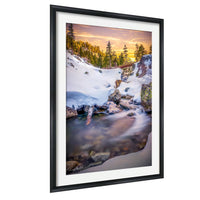 A framed photograph depicting a serene winter landscape with a snow-covered river flowing through a forested area, with a warm sunset sky in the background.